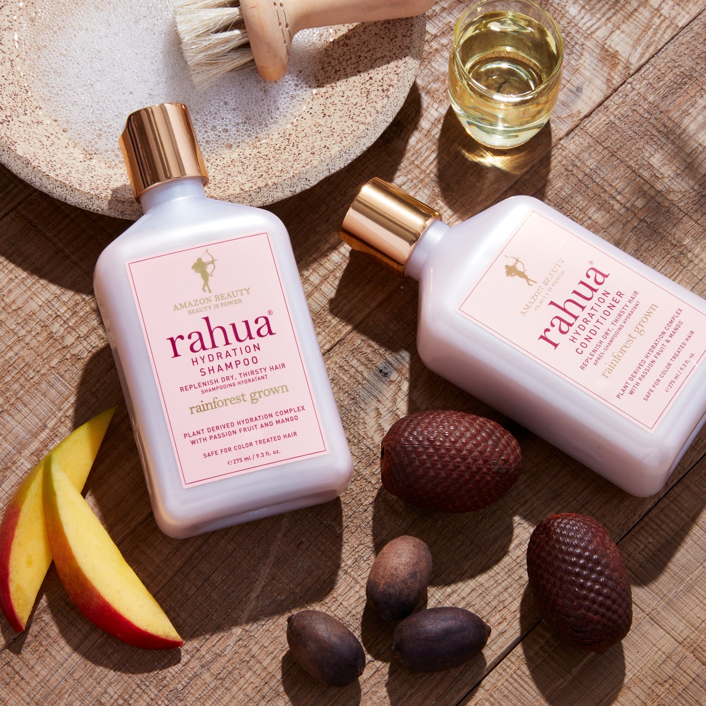 Two bottles of Rahua hair care products on a wooden surface with natural element featuring key ingredients like rahua oil morete oil and mango slices|variant:93-oz