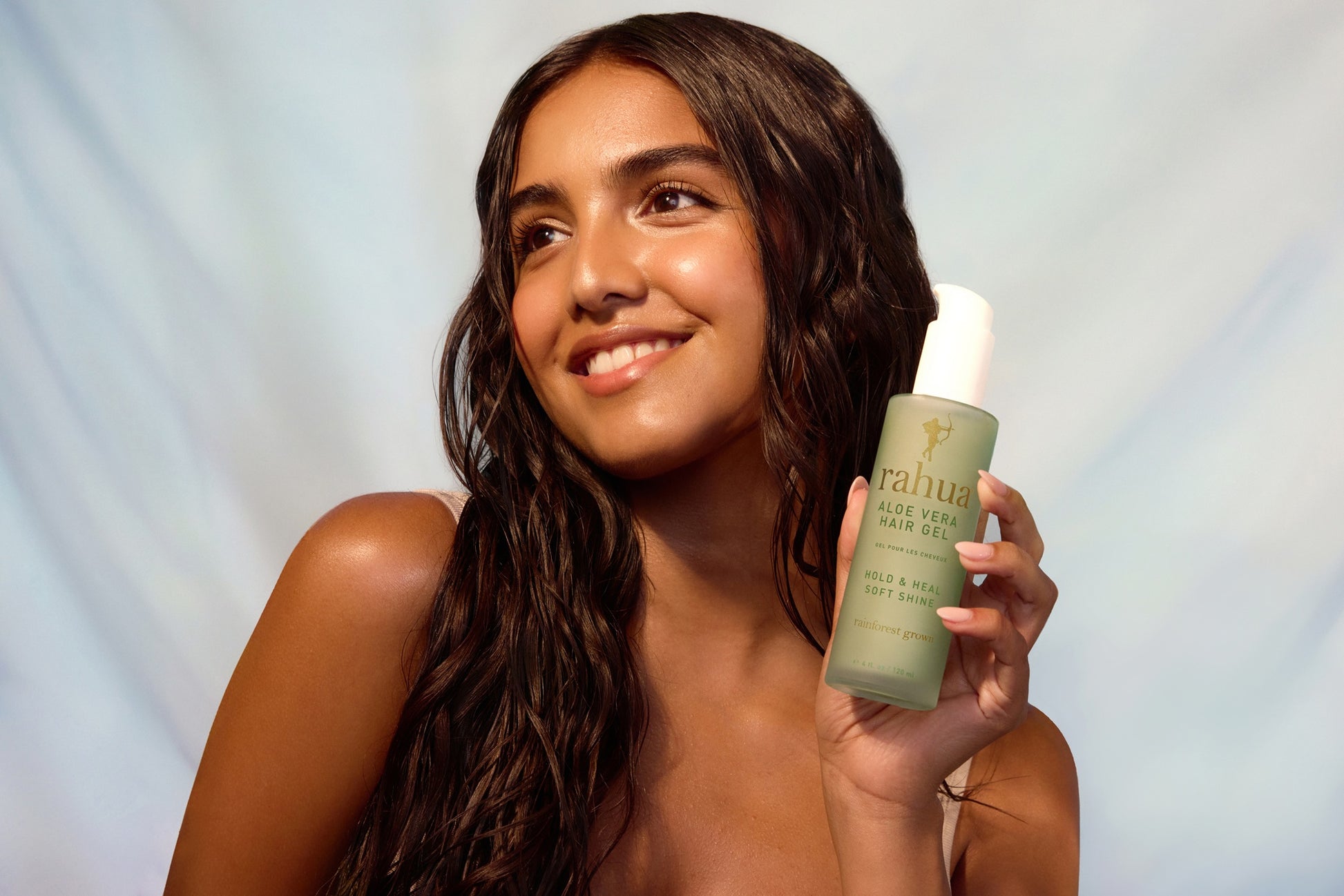 model with curly shiny hair in gentle waves holding a glass bottle of Rahua Aloe Vera Hair Gel set on a light blue grey gradient background|variant:4-oz