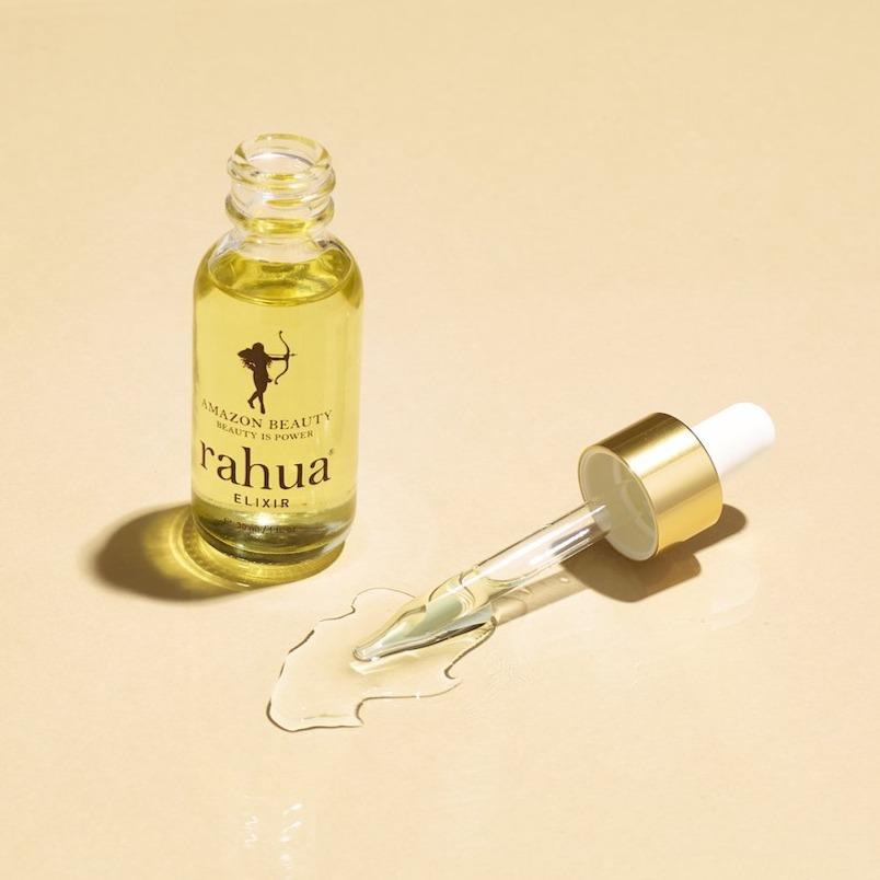 bottle of golden liquid in a glass bottle rahua elixir oil made of 100% rahua oil with a mini bottle next to it on a plain beige background with an orchid|variant:015-oz
